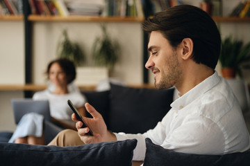 Aufkleber - Side view of young smiling businessman sitting on sofa with colleague on background while happily using cellphone in modern co-working space