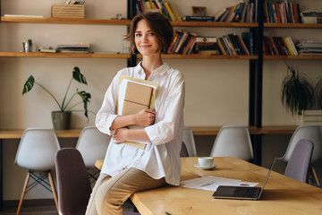Aufkleber - Young beautiful woman in white shirt happily looking in camera while sitting on desk with papers and laptop in modern office
