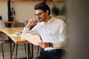 Aufkleber - Young attractive smiling businessman in eyeglasses happily reading papers while working in modern office