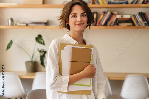 Fotomural Young beautiful smiling woman dreamily looking in camera with papers and notepad