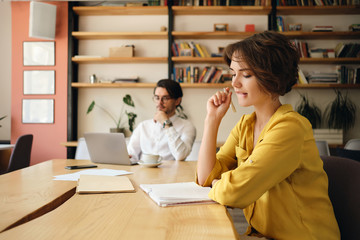 Aufkleber - Young attractive woman sitting at the table with notepad and dreamily working with colleague on background in modern office