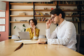 Aufkleber - Young handsome man in eyeglasses sitting at the table thoughtfully working with colleague on background in office
