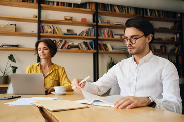 Aufkleber - Young serious man in eyeglasses sitting at the table thoughtfully working in modern office with colleague on background