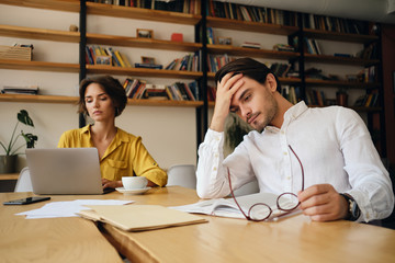 Aufkleber - Young tired man sitting at the table with notepad and eyeglasses while thoughtfully holding hand near head at work in office with colleague on background