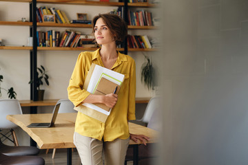 Aufkleber - Young beautiful woman in yellow shirt leaning on desk with notepad and papers in hand while thoughtfully looking aside in modern office