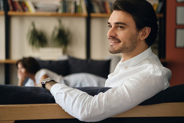 Aufkleber - Young attractive smiling businessman sitting on sofa with colleague while happily looking aside in modern co-working space