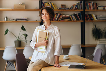 Aufkleber - Young attractive woman in white shirt happily looking in camera sitting on desk with papers and laptop in office