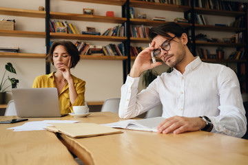 Aufkleber - Young tired man in eyeglasses sitting at the table leaning head on hand while thoughtfully working in office with colleague on background