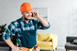 © LIGHTFIELD STUDIOS - cheerful worker in safety helmet talking on smartphone while standing with hand on hip