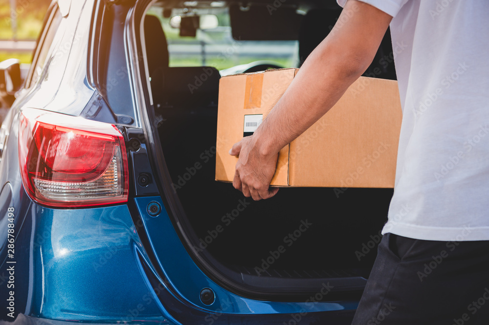 Delivery man is delivering cardboard box to customers via private car trunk door. People lifestyles and business occupation concept. Young male courier in casual clothes. Parcel move courier service - obrazy, fototapety, plakaty 