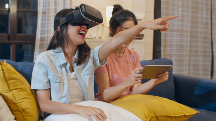 Wall Mural - Lesbian lgbt women couple using tablet at home, Asian female feeling happy using laptop and VR playing games together while lying sofa in living room in night. Lover celebrate holiday concept.