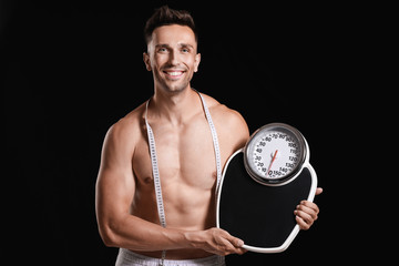 Handsome muscular man with measuring tape and scales on dark background. Weight loss concept