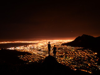 Two friends holding hands looking over Cape Town city lights from on top of Lion's Head at night, South Africa.