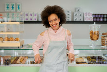 Beautiful Smiling Mixed Race Female Owner Of Pastry Shop Dressed In Apron Leaning On Counter In Shop.