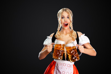 Young sexy oktoberfest girl waitress, wearing a traditional Bavarian or german dirndl, serving two big beer mugs with drink isolated on black background.