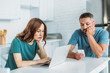 © LIGHTFIELD STUDIOS - thoughtful man using smartphone while sitting at kitchen table near wife using laptop
