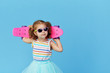 © oes - Cool little toddler girl in bright clothes and sunglasses carrying a skateboard over his shoulder and looking at the camera isolated on blue background