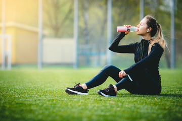 young pretty thirsty blonde Caucasian girl in suit resting after sport run in the football stadium sitting on grass drinking water and smiling enjoying and listening music with her wireless headphones