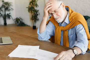 Portrait of stressed upset mature Caucasian businessman in formal clothes and glasses sitting in front of open laptop, studying documents, facing financial problems, holding hand on his bald head