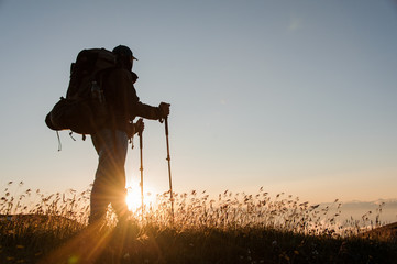 Man standing on the hill with hiking backpack and sticks looking at the sunset