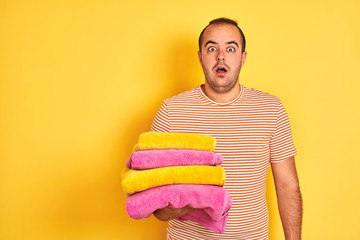 Sticker - Young shopkeeper man holding folded towels standing over isolated yellow background scared in shock with a surprise face, afraid and excited with fear expression