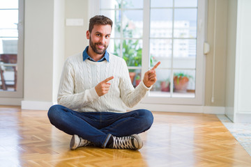 Wall Mural - Handsome man wearing casual sweater sitting on the floor at home smiling and looking at the camera pointing with two hands and fingers to the side.