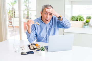 Canvas Print - Handsome business senior man eating delivery sushi while working using laptop stressed with hand on head, shocked with shame and surprise face, angry and frustrated. Fear and upset for mistake.