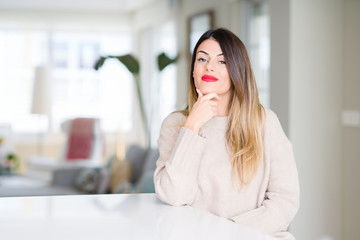 Sticker - Young beautiful woman wearing winter sweater at home looking confident at the camera with smile with crossed arms and hand raised on chin. Thinking positive.