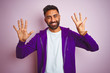 Young indian man wearing purple sweatshirt standing over isolated pink background showing and pointing up with fingers number nine while smiling confident and happy.