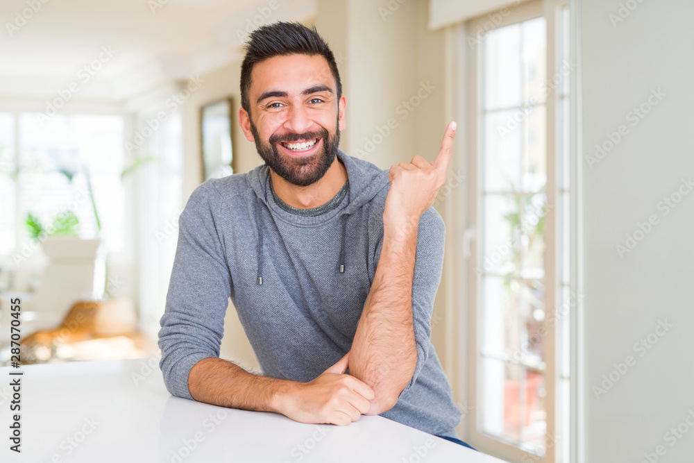 Handsome hispanic man wearing casual sweatshirt at home with a big smile on face, pointing with hand and finger to the side looking at the camera.