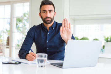 Poster - Handsome hispanic man working using computer and writing on a paper with open hand doing stop sign with serious and confident expression, defense gesture