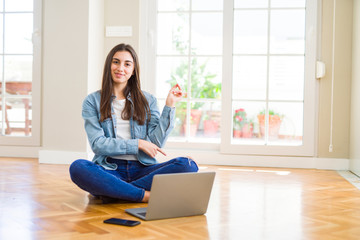 Poster - Beautiful young woman sitting on the floor with crossed legs using laptop with a big smile on face, pointing with hand and finger to the side looking at the camera.