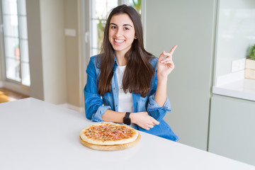 Sticker - Beautiful young woman eating homemade tasty pizza at the kitchen with a big smile on face, pointing with hand and finger to the side looking at the camera.