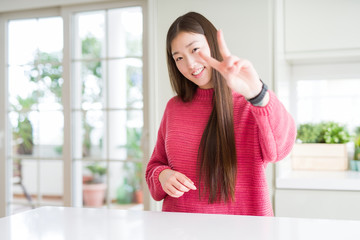 Poster - Beautiful Asian woman wearing pink sweater on white table smiling looking to the camera showing fingers doing victory sign. Number two.