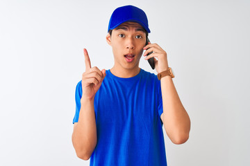 Wall Mural - Chinese deliveryman wearing cap talking on the smartphone over isolated white background surprised with an idea or question pointing finger with happy face, number one