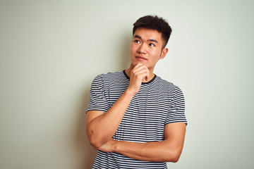 Wall Mural - Young asian chinese man wearing striped t-shirt standing over isolated white background with hand on chin thinking about question, pensive expression. Smiling with thoughtful face. Doubt concept.