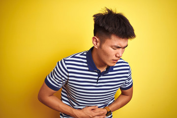 Sticker - Young asian chinese man wearing striped polo standing over isolated yellow background with hand on stomach because indigestion, painful illness feeling unwell. Ache concept.