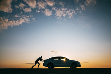 Wall Mural - Silhouette of man driver pushing his car along on an empty road after breakdown at sunset, copy space, side view. Road trip, trouble on the road 