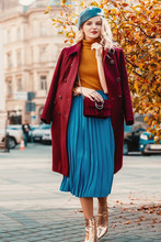 Street Style Autumn Fashion Concept: Full-length Portrait Of Elegant Lady Wearing Burgundy Color Woolen Coat, Light Blue Pleated Skirt, Beret, Metallic Color Ankle Boots, Holding Small Quilted Bag 