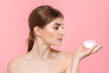 Wall Mural - Beautiful girl holding tube of cream to moisturize skin. Model looks at the camera isolated over pink background. Concept of beauty and health treatment.