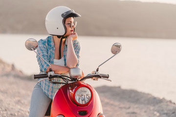 Wall Mural - Young pretty woman's portrait. Girl in white helmet and eyeglasses sitting on red retro scooter at nature background.