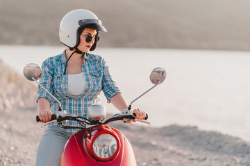 Wall Mural - Attractive girl's portrait. Lady in colorful clothes and sunglasses sitting on red retro moped at sunset background. Travel concept.