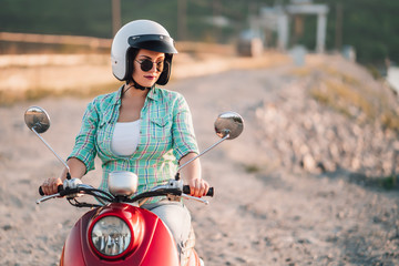 Wall Mural - Young pretty woman's portrait. Girl in white helmet and eyeglasses sitting on red retro scooter at nature background.