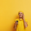 © WHstudio Leushin N - Glad male model calls friend via cellphone, holds mobile phone, enjoys conversation while drinking takeout coffee, wears yellow clothes, transparent glasses, poses indoor. People and lifestyle