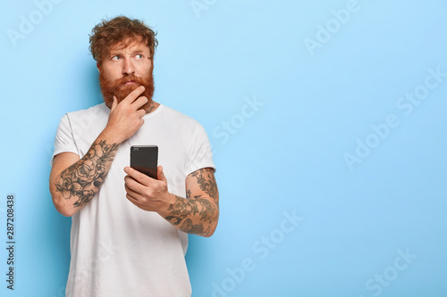 Horizontal shot of serious contemplative adult man touches thick red beard, holds mobile phone, browses newsfeed online, thinks over recent news, has tattooed arms, wears casual white t shirt