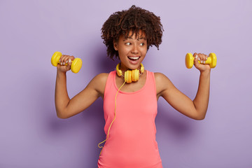 Pleased dark skinned model with curly hair, dressed in casual rosy t shirt, raises arms with dumbbells, trains muscles, listens music via headphones, isolated over purple background. People and sport