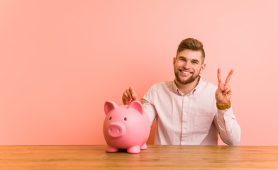 Young caucasian man sitting with a piggy bank showing number two with fingers.