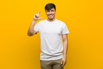 Wall Mural - Young hispanic casual man holding something little with forefingers, smiling and confident.