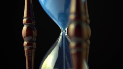 Wall Mural - Rotating hourglass clock time concept. Old vintage wood frame blue sand clock slowly spinning on dark background. Close up.