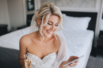 a beautiful, smiling blonde bride in a white dress is sitting on the bed and looking at the phone, r